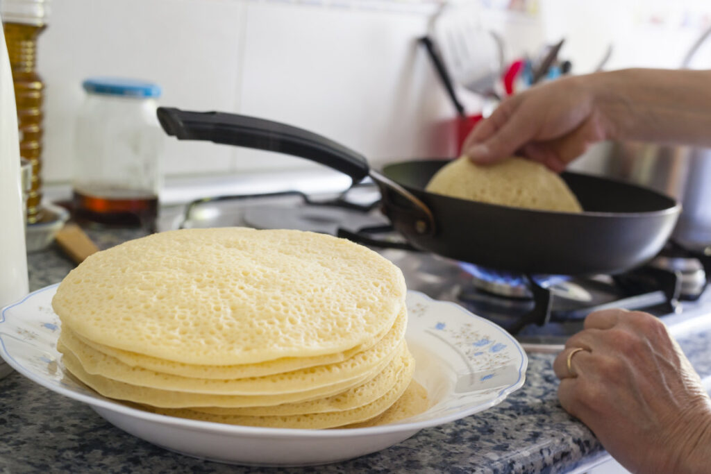 Hands of adult woman cooking baghrir on a pan with fire stoves on a home kitchen background with a pile of the typical moroccan pancakes on a big plate freshly cooked for breakfast.